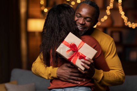 Téléchargez les photos : Happy birthday concept. Closeup of thankful african american handsome man with gift box hugging his wife and smiling at cozy home decorated with festive lights, copy space - en image libre de droit