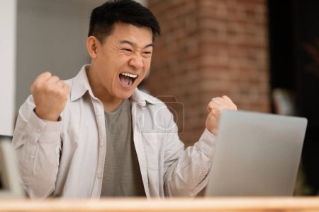 Téléchargez les photos : Happy mature asian male freelancer using laptop and shaking fists in joy, celebrating business success, sitting at table at home office. Successful freelance concept - en image libre de droit