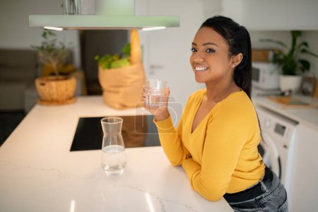 Téléchargez les photos : Happy young african american woman sit at table drinking water from glass in modern kitchen interior with food package, free space. Aqua balance, slimming and weight loss, diet, body and health care - en image libre de droit