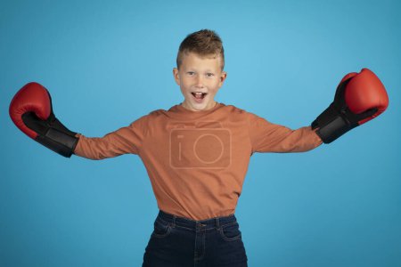 Téléchargez les photos : Portrait Of Preteen Caucasian Boy Wearing Boxing Gloves Posing Over Blue Background, Male Child Spreading Hands And Looking At Camera, Excited Kid Enjoying Mai Sport Training, Copy Space - en image libre de droit
