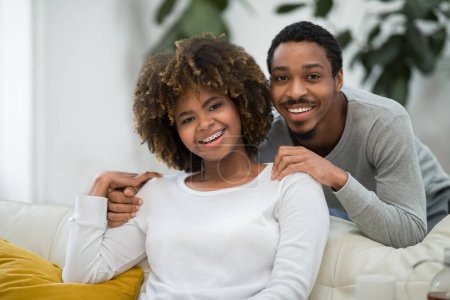 Photo for Portrait of beautiful happy loving black couple bonding at home, happy handsome millennial man hugging his pretty curly wife from behind, smiling at camera, living room interior, copy space - Royalty Free Image