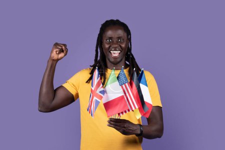 Foto de Emotional handsome long-haired millennial african american guy in yellow t-shirt holding diverse flags of various countries and gesturing, purple studio background. Education abroad, immigration - Imagen libre de derechos
