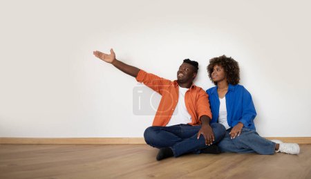 Foto de Happy Young African American Spouses Sitting On Floor Near White Wall And Pointing Aside, Smiling Black Couple Planning Design In Their New Home After Moving And Demonstrating Free Copy Space - Imagen libre de derechos