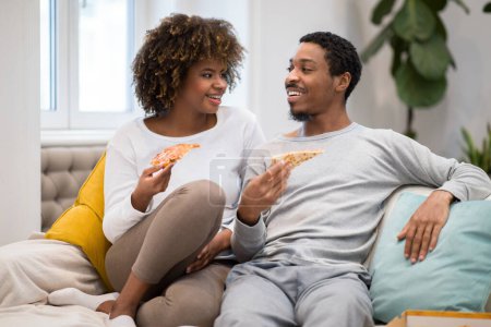 Téléchargez les photos : Happy beautiful millennial black lovers sitting on couch, eating pizza, having conversation and smiling, loving african american man and woman spending weekend together at home, copy space - en image libre de droit