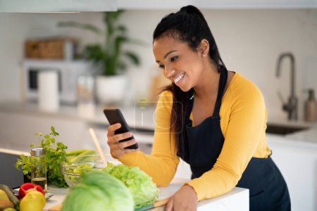 Photo for Smiling young african american woman in apron prepare salad, watch video, typing on smartphone at table with fresh vegetables in kitchen interior. App for cook healthy food at home, chef and blog - Royalty Free Image