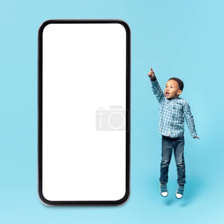 Photo for Educational kids mobile app. Surprised black little boy pointing aside and jumping near big smartphone with blank screen, blue studio background, mockup - Royalty Free Image