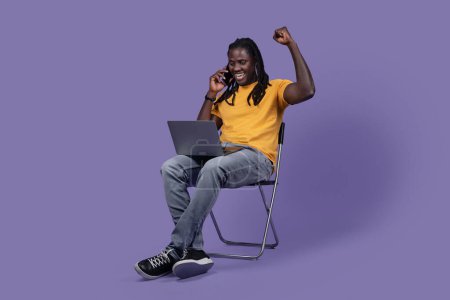 Photo for Emotional handsome long-haired young black man in casual entrepreneur sitting on chair over purple studio background, using modern laptop and talking on phone, working online, copy space - Royalty Free Image