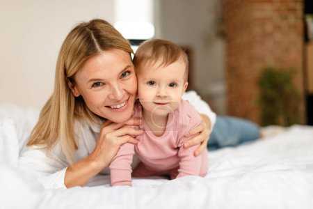 Téléchargez les photos : Happy motherhood. Mommy playing with adorable toddler baby crawling on bed, smiling together at camera, bedroom interior. Mother enjoying child care routine during maternity leave - en image libre de droit