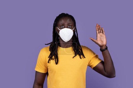 Photo for Concerned african american young man with protective mask of coronavirus disease showing hand in volunteer sign, looking at camera with enthusiastic expression, purple studio background - Royalty Free Image
