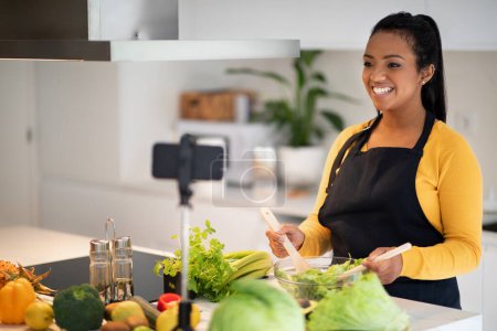 Téléchargez les photos : Happy young african american woman in apron prepare salad at table with fresh vegetables, shoot video on phone in modern kitchen interior. Blogger cook healthy eat at home, diet, food blog at home - en image libre de droit