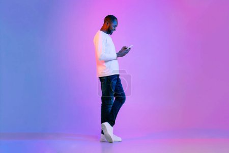 Téléchargez les photos : Cheerful smiling handsome middle aged black man in casual posing on neon light background, using cell phone, checking newest mobile app, surfing on Internet, full length studio shot, copy space - en image libre de droit