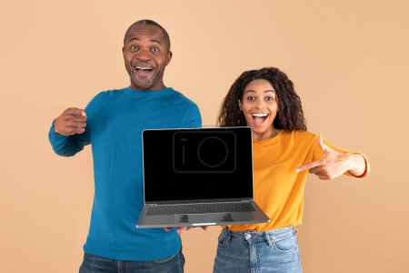 Téléchargez les photos : Cool website. Happy black adult couple pointing at laptop with blank screen, middle aged man and young lady pointing at copy space for advertisement, posing over peach background, mockup - en image libre de droit