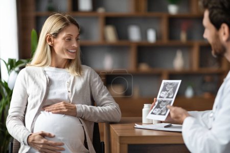 Foto de Gynecologist Doctor Showing Baby Sonography Image To Young Pregnant Female During Meeting In Clinic, Smiling Reproductive Endocrinologist Consulting Happy Expectant Woman In Hospital, Free Space - Imagen libre de derechos