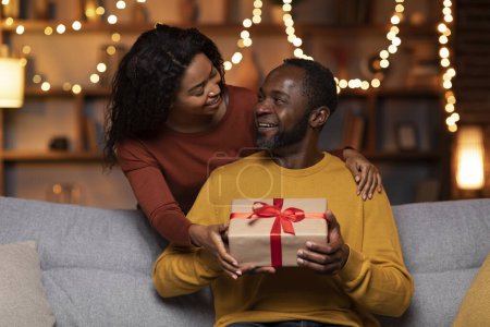 Téléchargez les photos : Happy anniversary my love. Cheerful pretty young african american woman greeting her beloved spouse or boyfriend, hugging man from behind and giving gift box, festive home interior, copy space - en image libre de droit