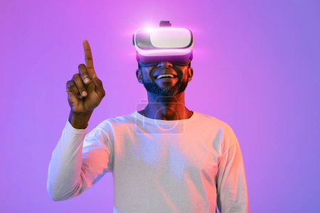 Photo for Joyful middle aged african american man in white touching something invisible with finger and smiling, trying augmented reality, using modern VR glasses over neon studio background - Royalty Free Image