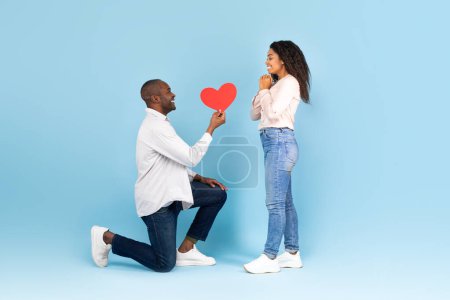 Foto de Romantic african american man on his knees giving red heart shaped card to excited black woman, posing together on blue studio background, side view - Imagen libre de derechos