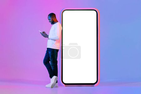 Téléchargez les photos : Happy handsome middle aged african american man in casual outfit posing with huge phone with white blank screen on neon light studio background, using phone, chatting with ladies on dating app, mockup - en image libre de droit