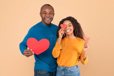Téléchargez les photos : Love is in the air. Romantic black couple with red paper hearts in hands posing over peach studio background, middle aged man and young woman smiling at camera - en image libre de droit