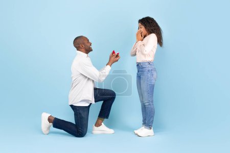 Foto de Marry me. Black middle aged man holding giving open box with engagement ring to excited young woman, asking her to be his wife during romantic date standing on one knee, blue studio background - Imagen libre de derechos
