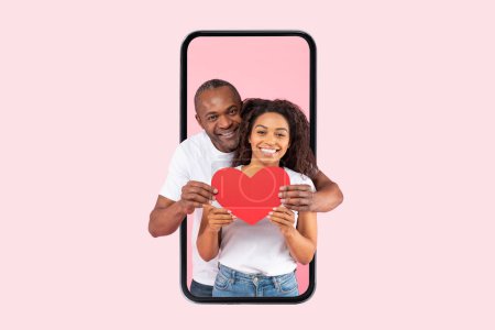 Foto de Love and care. Happy african american spouses hugging holding red paper card heart standing inside smartphone screen on pink studio background. St. Valentines Day - Imagen libre de derechos