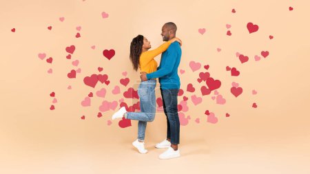 Photo for Loving black couple hugging and smiling at each other, standing on peach studio background with red flying hearts, side view, panorama. St. Valentines Day - Royalty Free Image
