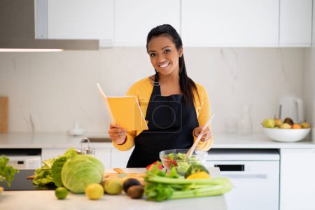 Photo for Cheerful young african american female in apron preparing salad and reading recipe book at table with fresh vegetables in modern kitchen interior. Homemade food, diet, cooking healthy eat at home - Royalty Free Image