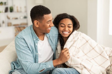 Téléchargez les photos : Cheerful young african american guy wraps lady in blanket, couple enjoys warm together, sitting on sofa in living room interior. Care and support in winter, romance, love and relationships at home - en image libre de droit