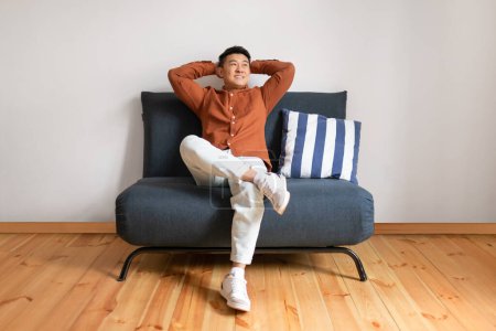 Foto de Lazy weekend. Happy mature asian man relaxing with hands behind head, sitting on sofa over white wall. Relaxation and comfort concept - Imagen libre de derechos