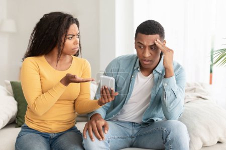 Téléchargez les photos : Unhappy jealous angry young african american wife show smartphone to sad confused husband accuses of treason in home interior. Addiction from gadget and online games, relationship problems, cheating - en image libre de droit