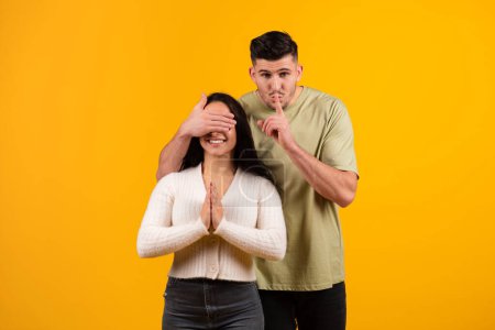 Foto de Cheerful millennial arabic guy closes eyes to amazed wife, makes shhh sign, preparing surprise for anniversary, isolated on orange background, studio. Congratulations to holiday, make wish at birthday - Imagen libre de derechos