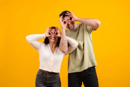 Téléchargez les photos : Happy millennial middle eastern male and female have fun doing glasses with hands, enjoy free time together, isolated on orange background, studio. Entertainment and relationships, love and romantic - en image libre de droit