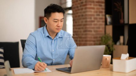 Foto de Confident middle aged asian man making notes, working on laptop in home office interior, panorama, free space. New normal, remote education, freelance due covid-19 quarantine - Imagen libre de derechos