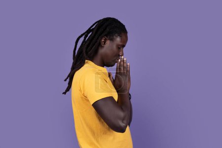 Téléchargez les photos : Side view of calm spiritual handsome black guy praying with closed eyes. Serious peaceful man with joining hands meditating. Isolated on purple studio background. Belief concept, copy space - en image libre de droit
