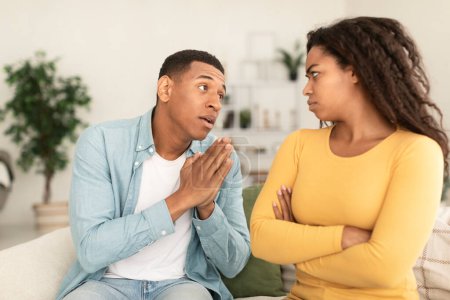 Foto de Sad young african american man asking for forgiveness from angry offended female in living room interior, free space. Apologize, sorry, relationship problems, people emotions after quarrel at home - Imagen libre de derechos
