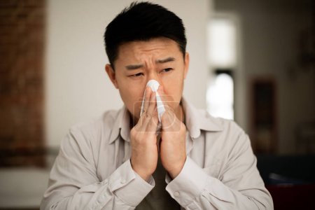 Photo for Unhappy middle aged asian man suffering from fever and flu, blowing nose in napkin, sitting in living room interior. Covid-19 lockdown, treatment of illness, cold and runny - Royalty Free Image