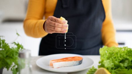 Foto de Young african american woman in apron pours lemon juice, cooks fish at table with fresh vegetables in kitchen interior, cropped. Job of professional chef in restaurant, diet, cook healthy food at home - Imagen libre de derechos