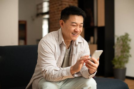 Happy korean middle aged man chatting on smartphone, watching video or surfing internet while resting on sofa in living room. Free time for blog at home, new app, advertisement and offer
