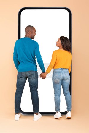 Téléchargez les photos : Happy black couple standing near huge phone with empty screen, standing back to camera, holding hands and smiling at each other. Great mobile app concept - en image libre de droit