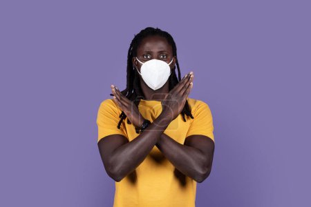 Foto de Young black guy with long braids in yellow t-shirt and protective medical mask gesturing stop on purple studio background, copy space. Stay home while pandemic concept - Imagen libre de derechos