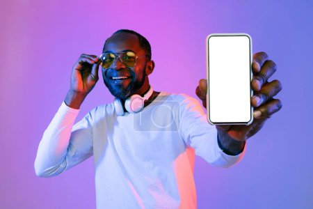 Téléchargez les photos : Stylish handsome cheerful mature black man in white showing modern smartphone with white empty screen, wearing yellow sunglasses and wireless headphones, recommending nice app in neon light, mockup - en image libre de droit