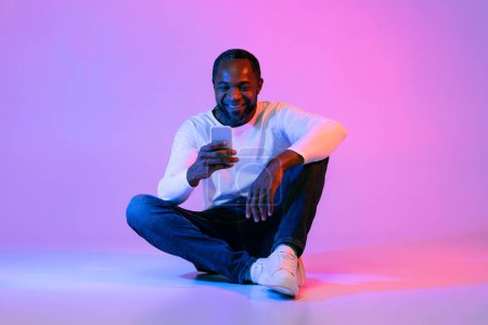 Photo for Happy handsome middle aged black man in casual outfit sitting on floor, using modern smartphone over neon studio background, gambling on Internet, chatting with ladies, copy space - Royalty Free Image