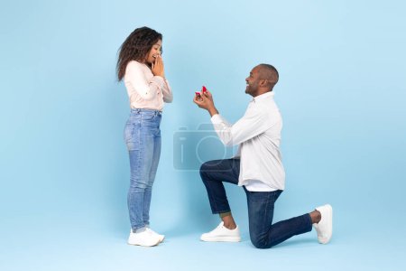 Téléchargez les photos : Loving black man standing on one knee and offering engagement ring to his beloved young woman on blue studio background, side view. Middle aged male making proposal to sweetheart - en image libre de droit