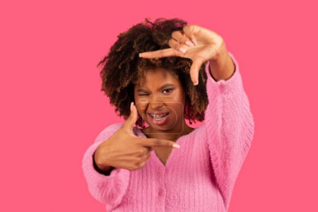 Téléchargez les photos : Happy young african american curly woman with braces show sign, make frame with hands, look through it, closing one eye with imaginary camera isolated on pink background. Creative and photography - en image libre de droit
