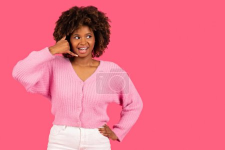 Téléchargez les photos : Happy pretty young african american curly woman with braces calling at imaginary phone, enjoy talk, look at free space isolated on pink background, studio. Gossip, good news, communication remotely - en image libre de droit
