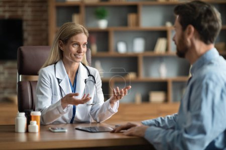 Foto de Smiling Doctor Woman In Uniform Consulting Male Patient During Meeting In Clinic, Therapist Lady Wearing White Coat And Stethoscope Sitting At Desk And Talking To Young Man, Explaining Treatment - Imagen libre de derechos