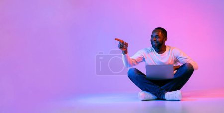 Téléchargez les photos : Smiling middle aged handsome black man in casual outfit digital nomad sitting on floor over neon light studio background, using laptop, pointing at copy space, recommending nice website, panorama - en image libre de droit
