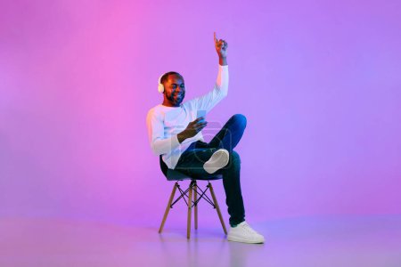 Téléchargez les photos : Cool mature black man in casual sitting on chair over neon light studio background, listening to music and moving his hands, using phone and wireless headphones, enjoying stereo sound, copy space - en image libre de droit