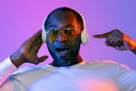 Photo for Amazed shocked middle aged handsome bearded black man in white wearing yellow sunglasses using modern stereo wireless headphones, pointing at gadget, neon studio background, closeup - Royalty Free Image