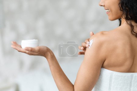 Foto de Smiling middle eastern millennial curly woman in towel holding jar, applies cream on her shoulder in bedroom interior with copy space. Beauty care and skin moisturizing, treatments at home, spa day - Imagen libre de derechos