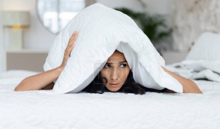 Photo for Sad muslim millennial curly female woke up, lies on white bed, covers head with pillow, suffers from depression and noisy neighbors in bedroom interior. Sleep problems, insomnia at night alone at home - Royalty Free Image